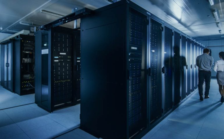 Shot of an IT Admin with a Laptop Computer and Young Technician Colleague Walking Next to Server Racks in Data Center. Running Diagnostics or Maintenance.